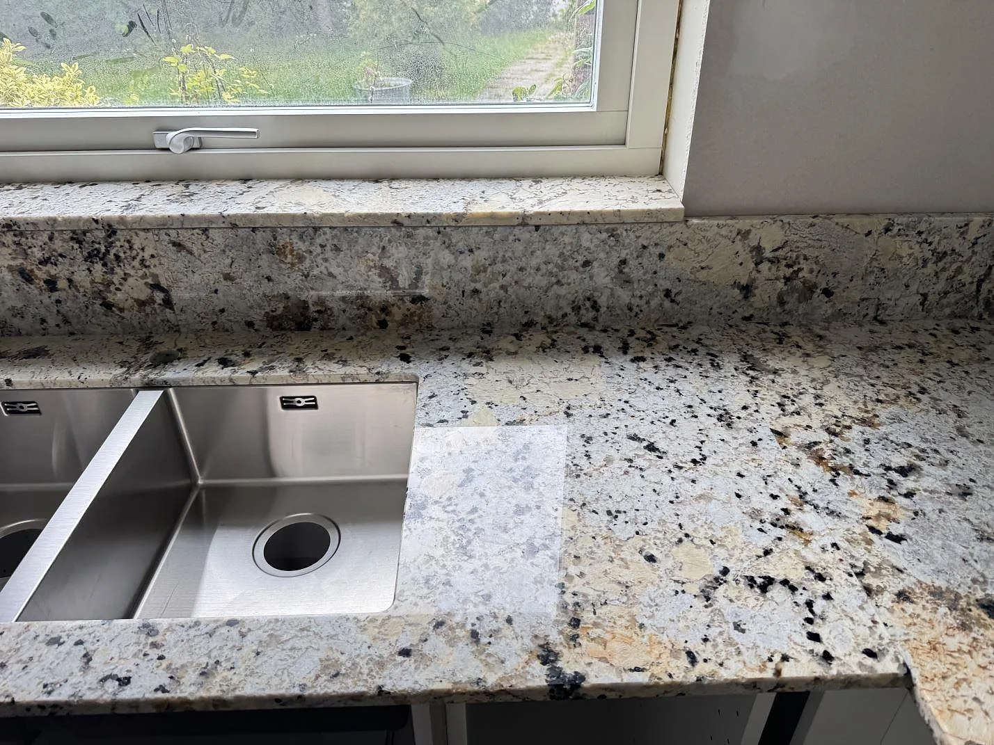 Polerad köksbänkskiva i Alaskan white granit vid fönster och rostfri diskho i Kristianstad.