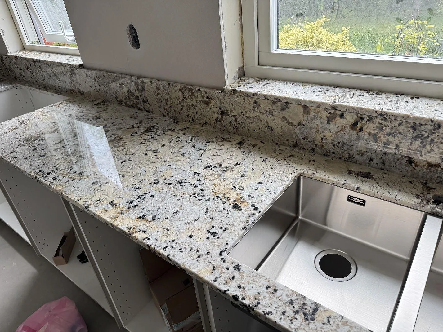 Köksbänkskiva i polerad Alaskan white granit med stänkkant, fönsterbänk och rostfri diskho i ett kök i Kristianstad.