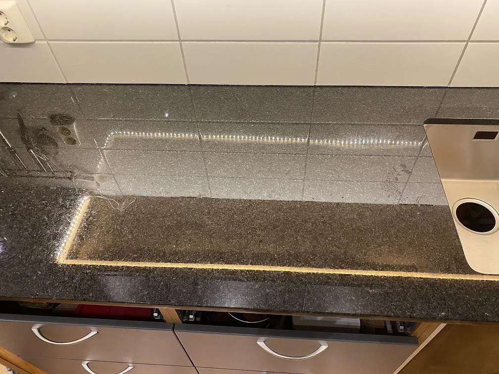 Lång bänkrad i polerad Angola Black granit med mörk stenyta, vit kakelvägg och diskmaskin vid sidan i Hässleholm.