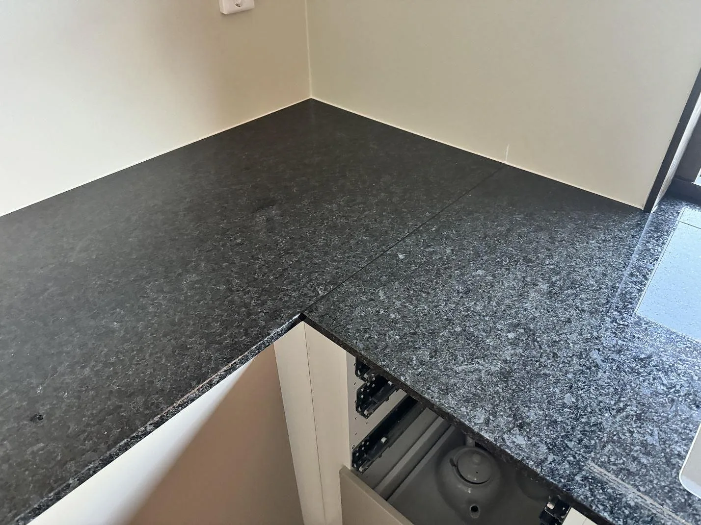 Hörnparti av köksbänkskiva i polerad Angola Black granit, mörk stenyta med ljus vägg och öppna stommar i HJÄRUP.