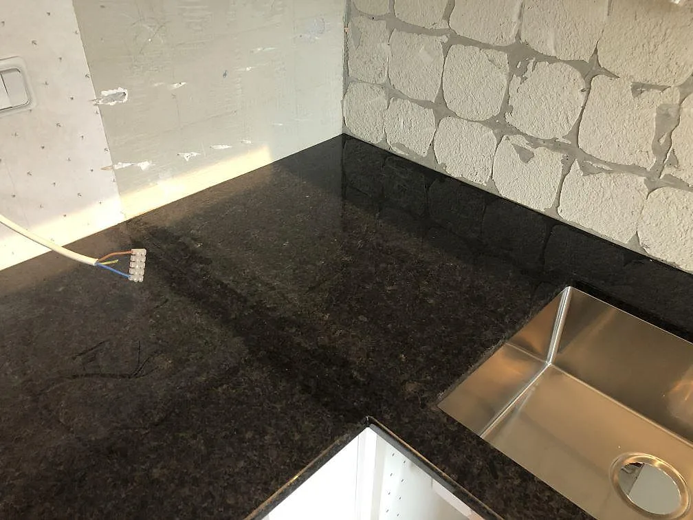 Hörn av köksbänkskiva i polerad svart Angola Black-granit med uppvikt stänkkant och underlimmad rostfri diskho i ett kök i Malmö.