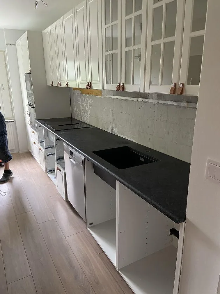 Köksbänkskiva och L formad bänk i Angola Black granit kring vita skåp med kopparfärgade beslag, fotograferat under montering i ett kök i Malmö.