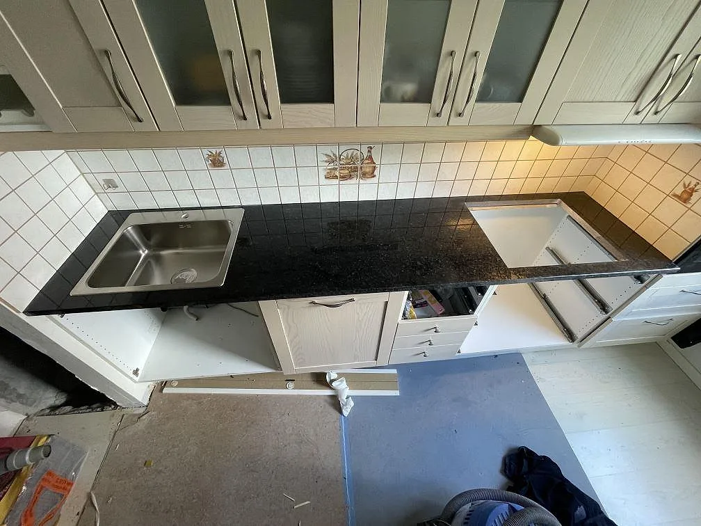 Köksbänkskiva i svart granit Angola Black, polerad, 30 mm, med underlimmad rostfri diskho och förberedd håltagning för häll mot vitt kakel i ett kök i Solna.