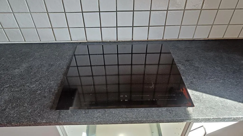 Svart induktionshäll i en matt Angola Black granitbänkskiva mot vitt kvadratiskt kakel i Staffanstorp.