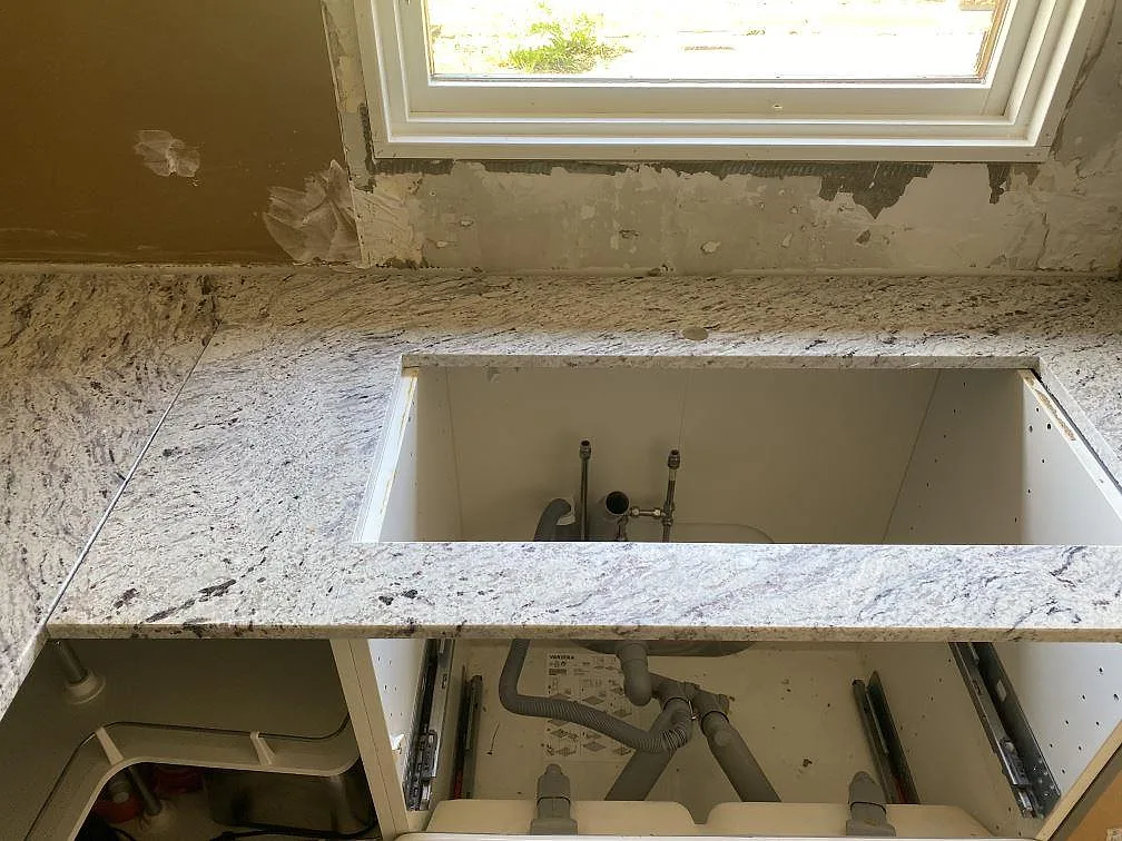 Polerad Antique White granit som köksbänkskiva under fönster, med diskhoöppning och öppna köksstommar i Älmhult.