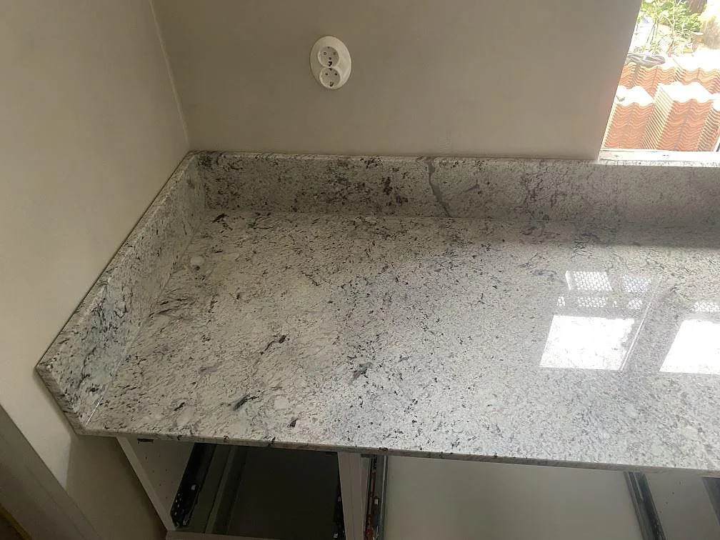 Köksbänkskiva i Antique White-granit, polerad, med uppvikt stänkkant och dagsljus som speglas i ytan i ett kök i Borrby.