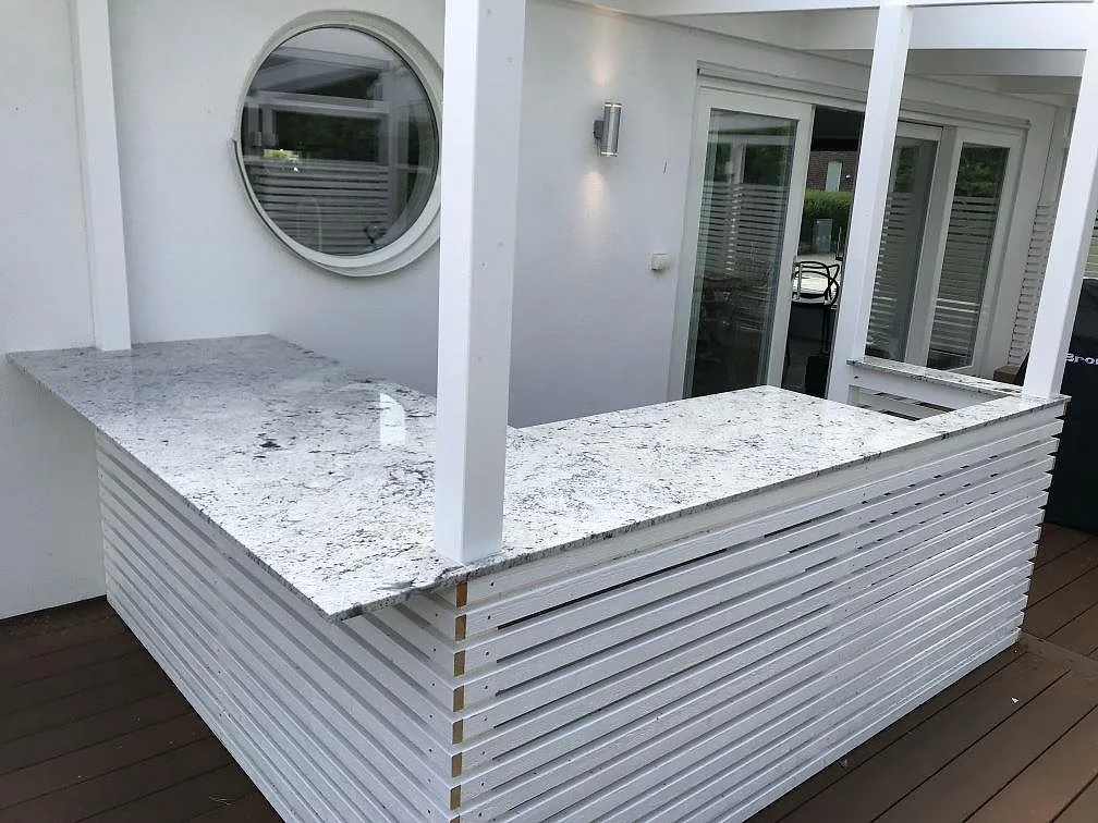 Köksbänkskiva i vit Antique White-granit, polerad, monterad som vinklad utomhusbänk på vita spjälstommar på ett trädäck i Dösjebro.