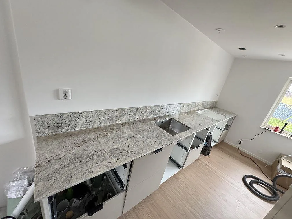 Köksbänkskiva i polerad Antique White granit med uppvikt stänkkant, rostfri diskho och hälluttag i ett kök under montering i Eslöv.
