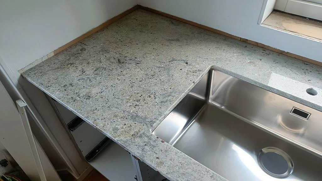 Närbild på polerad Antique White-granit med underlimmad rostfri diskho och kranhål, som visar stenens grå ådring och bruna inslag, Frillesås.