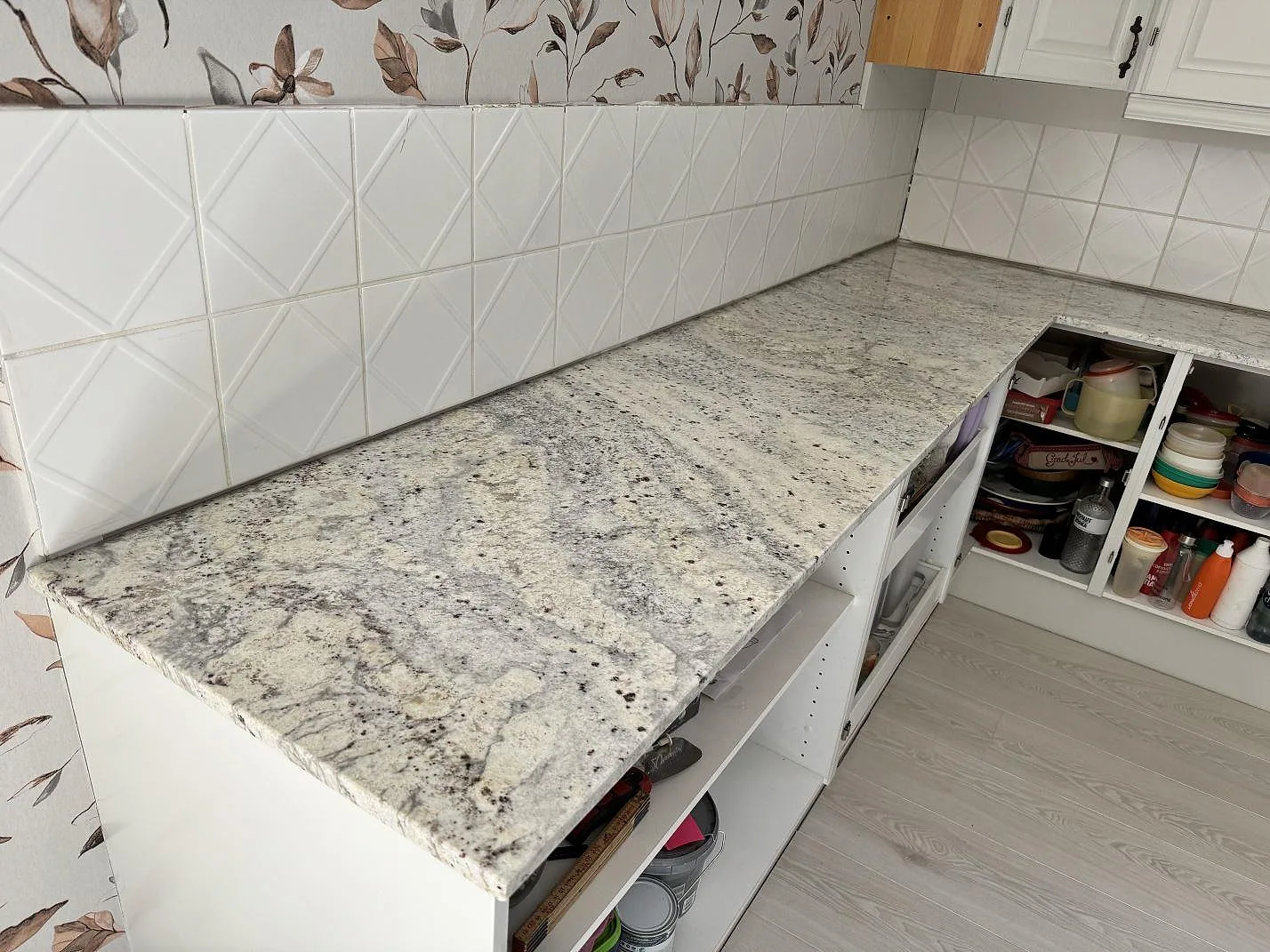 Lång köksbänkskiva i polerad Antique White granit med vit kakelvägg och blommig tapet i Gärds Köpinge.