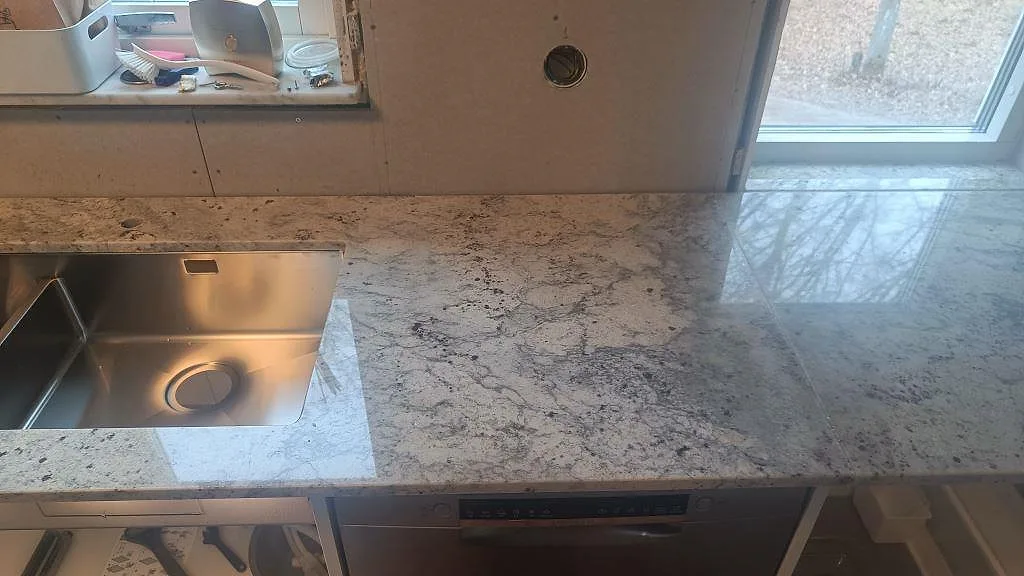 Detalj av polerad Antique White-granit kring rostfri diskho och hällzon i ett kök i Hörby.