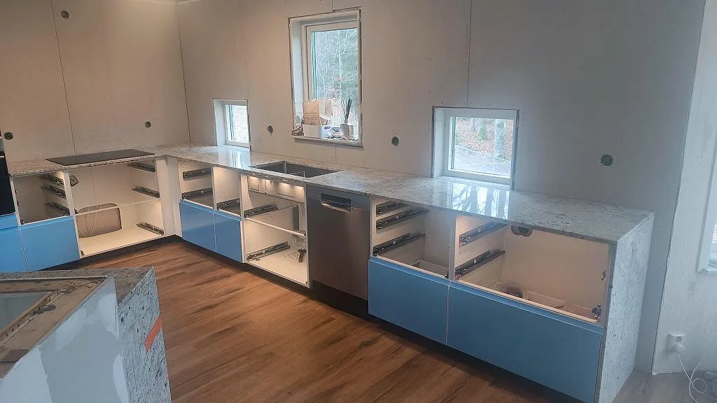 Köksbänkskiva i polerad Antique White-granit med halvö, diskho och blå frontpartier i ett kök i Hörby.
