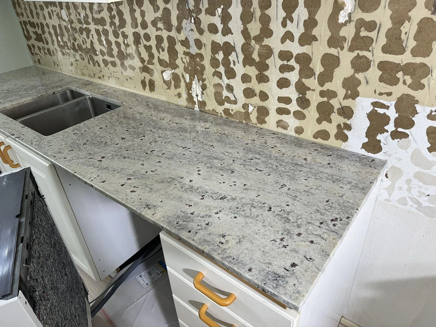 Köksbänkskiva i Antique White granit, polerad, längs en vägg med öppna stommar och ofärdig stänkyta i ett kök i Hörby.