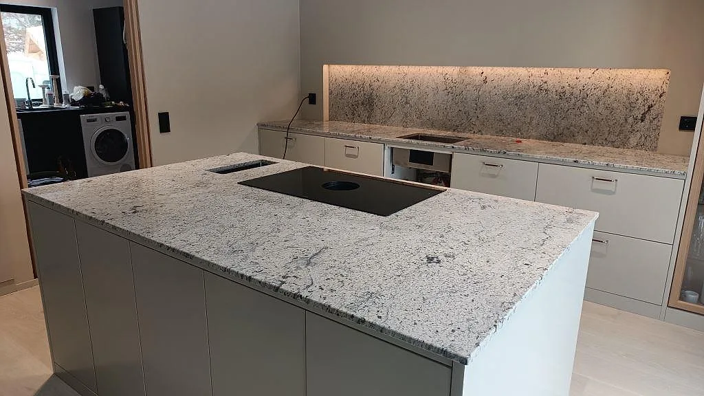Köksbänkskiva och köksö i vit Antique White-granit med induktionshäll och bakbelyst stänkvägg i ett kök i Kungsbacka.