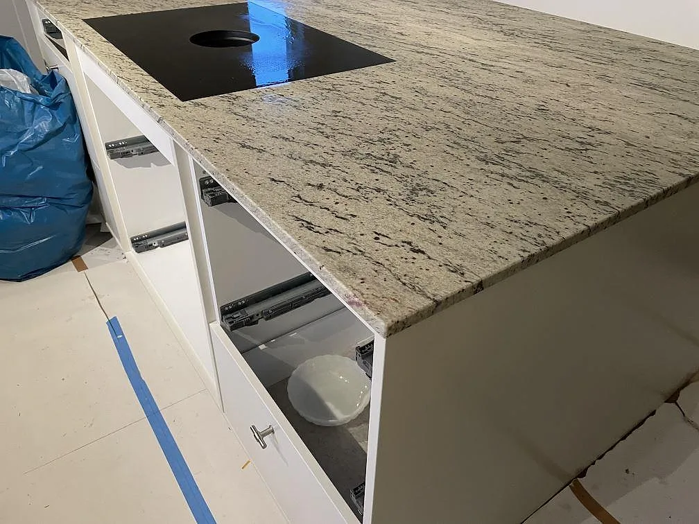 Stor arbetsyta i matt Antique White granit med svart induktionshäll och öppna ljusgrå stommar under montering i Landvetter.