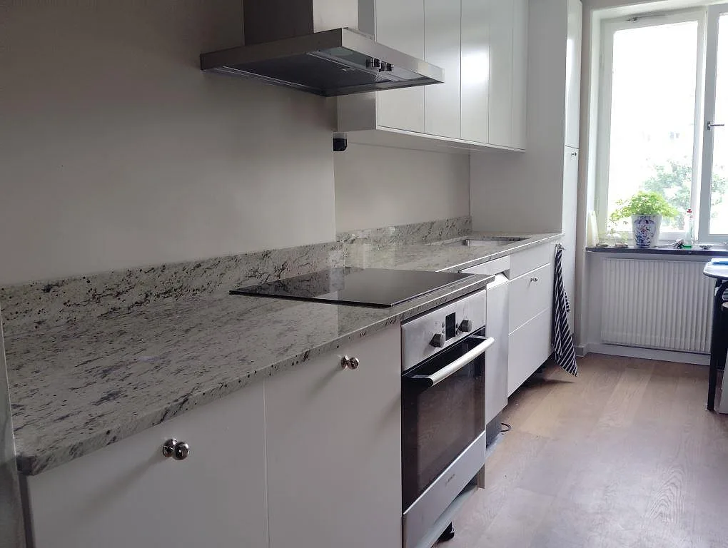 Polerad köksbänkskiva i Antique White granit med stänkkant, fläktkåpa och ljusa skåp i ett smalt kök i Lidingö.