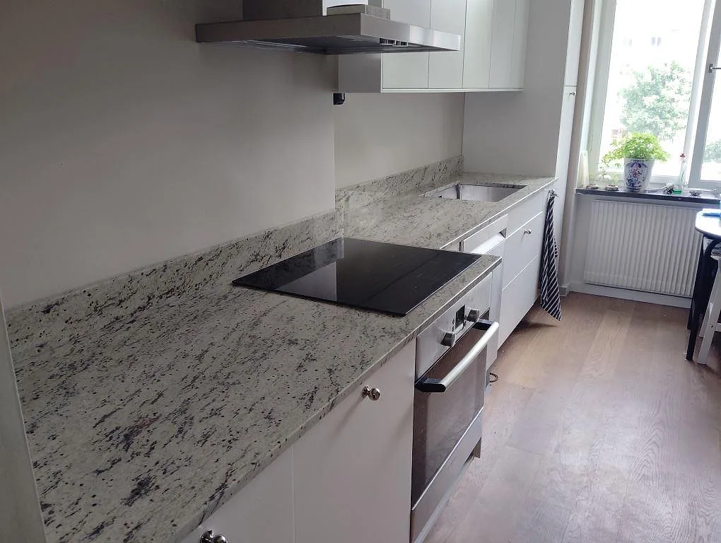 Köksbänkskiva i polerad Antique White granit med låg stänkkant, spiszon, diskhozon och fönster i ett kök i Lidingö.