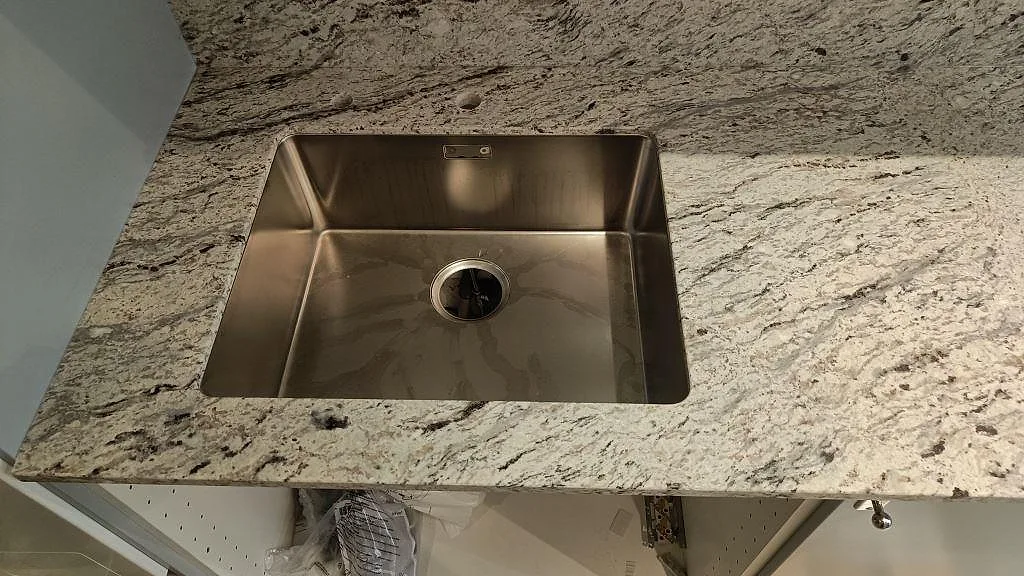 Närbild av rostfri diskho monterad under matt Antique White granit i ett kök under montering i Malmö.