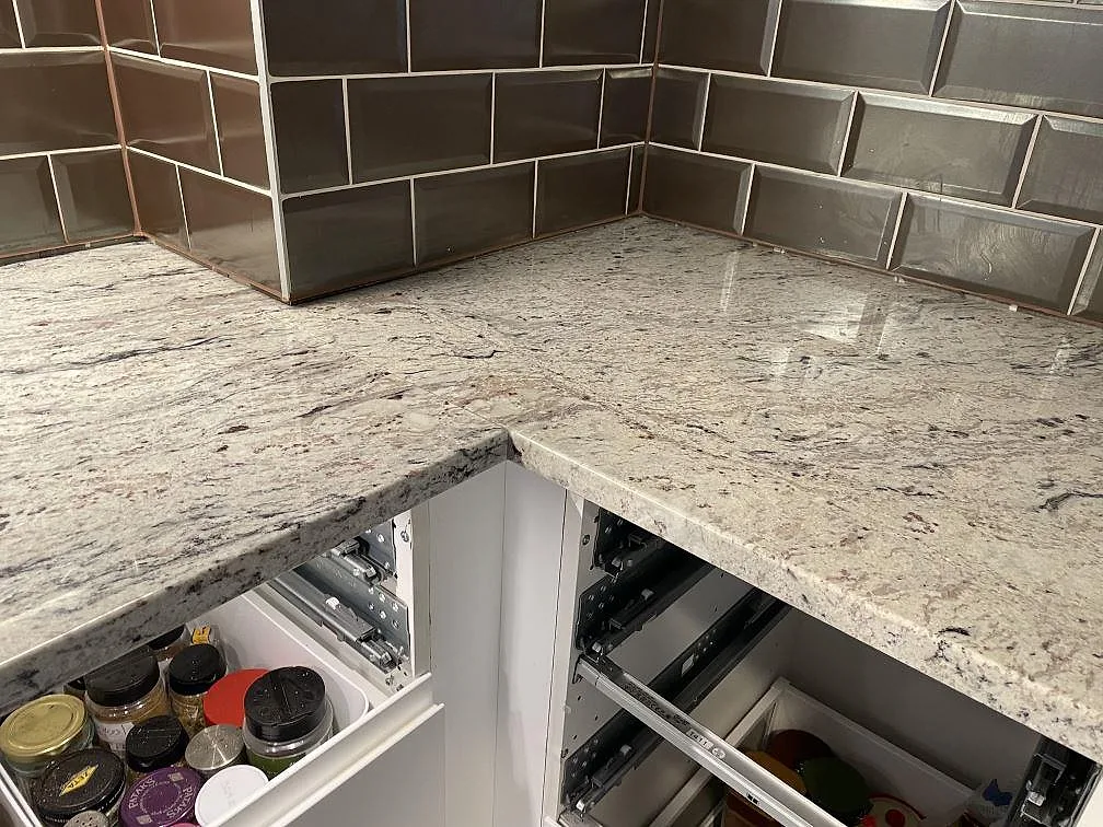 Hörnparti av L-formad köksbänkskiva i polerad Antique White granit mot brungrått kakel i Malmö.