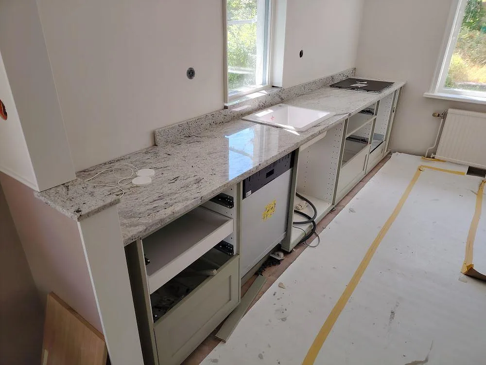Köksbänkskiva i polerad Antique White granit med låg stänkkant och öppna köksstommar under montering i Täby.