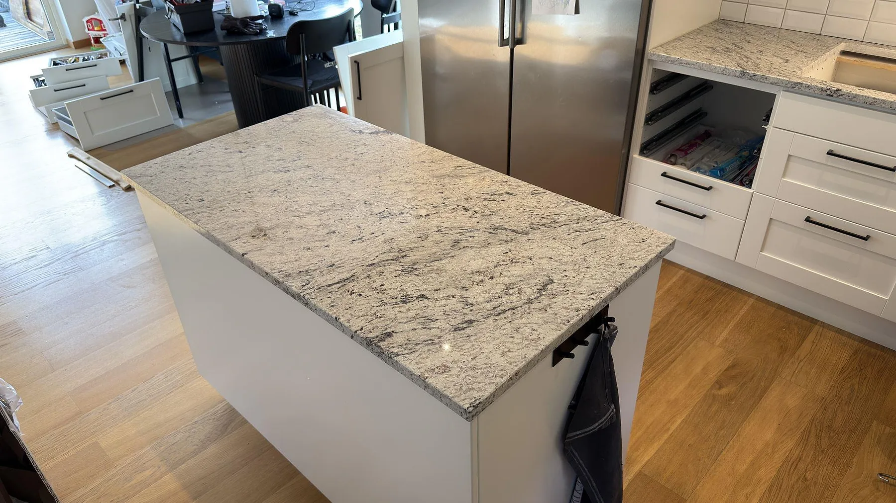 Köksbänkskiva i Antique White granit, polerad, på en fristående köksö i ett kök i Täby med vita luckor och svarta handtag.