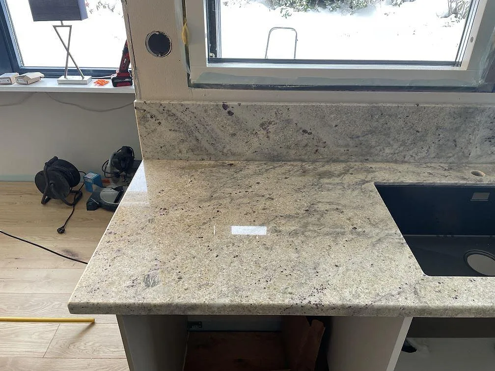 Köksbänkskiva i Antique White granit, polerad, med uppvikt stänkkant och underlimmad diskho framför fönster i ett kök i Trollhättan.