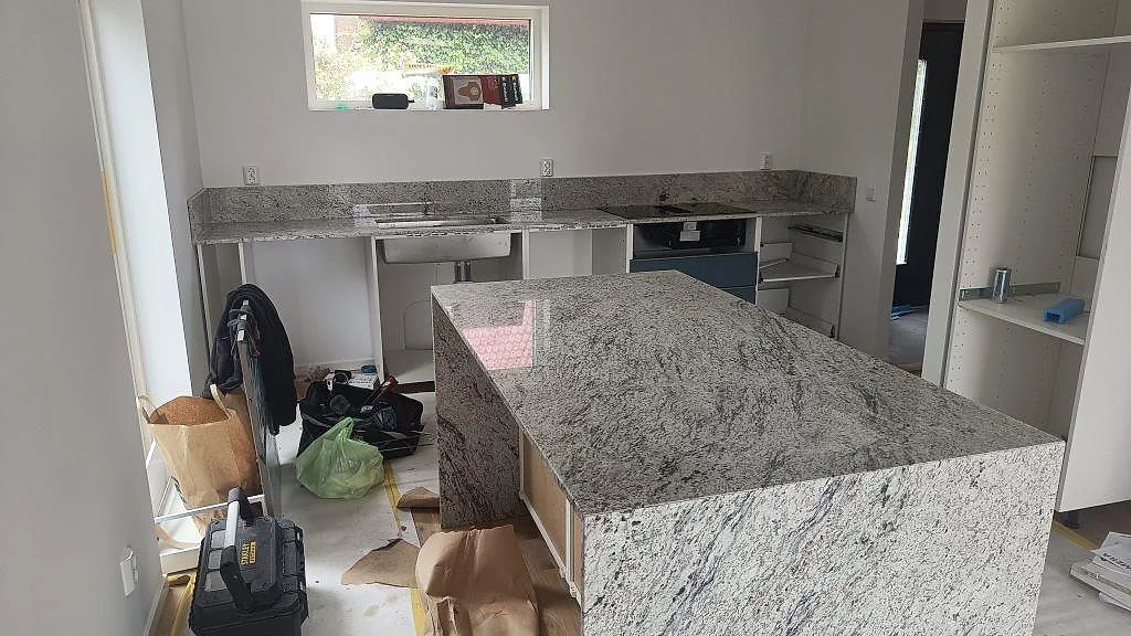 Köksbänkskiva och stor köksö i polerad Antique White granit med uppvikt stänkskydd i ett kök under montering i Trollhättan.