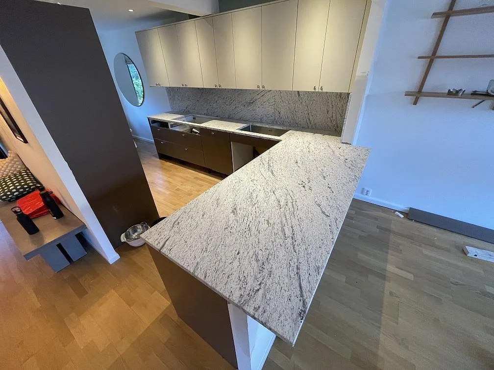 Köksbänkskiva i Antique White granit, matt yta, i ett L-format kök i Tyresö under montering med bakre bänkrad och stänkskydd i samma ljusa sten.