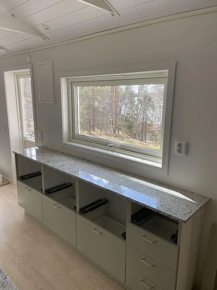 Köksbänkskiva i krämvit Antique White-granit längs en fönstervägg med köksstommar i Växjö.