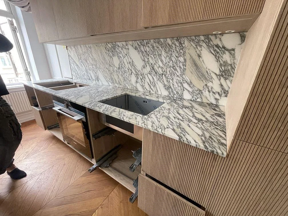 L-format kök i Malmö med matt Arabescato marmorbänkskiva, heltäckande marmorstänkvägg, ribbad träinredning och rostfri underlimmad diskho under montering.