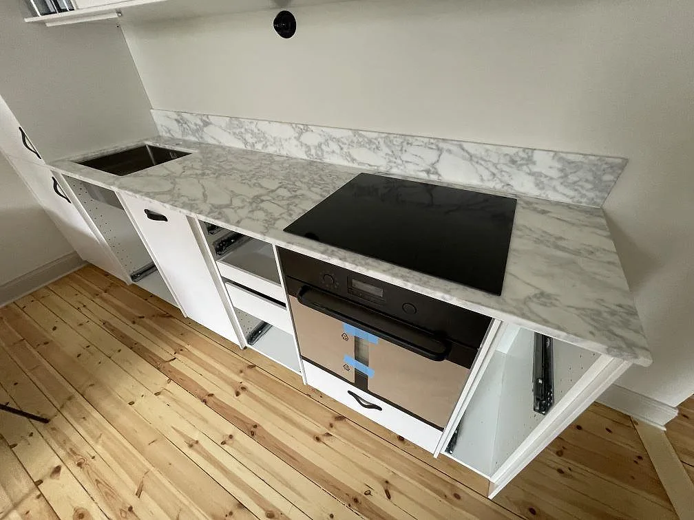 Köksbänkskiva i matt Arabescato-marmor med svart häll, diskhoöppning och låg stänkkant i ett kök i Stockholm.