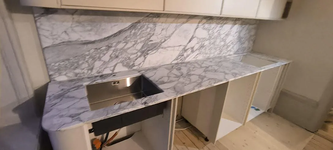 Köksbänkskiva i vit och grå Arabescato-marmor, matt, med matchande stänkskydd och diskhoöppning i ett kök i Stockholm.