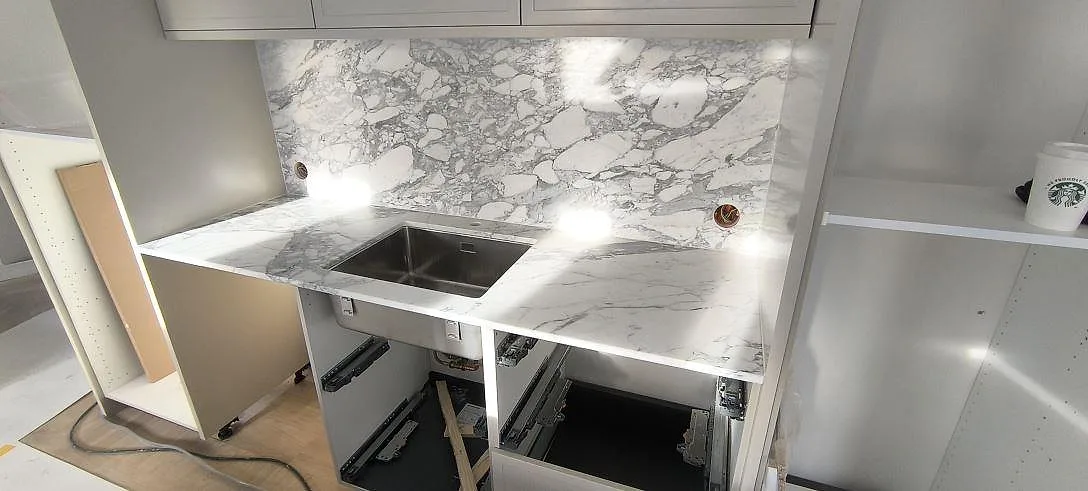 Köksbänkskiva i Arabescato marmor med underlimmad rostfri diskho och heltäckande stänkvägg i samma marmor bakom väggbänken i ett kök i Stockholm.