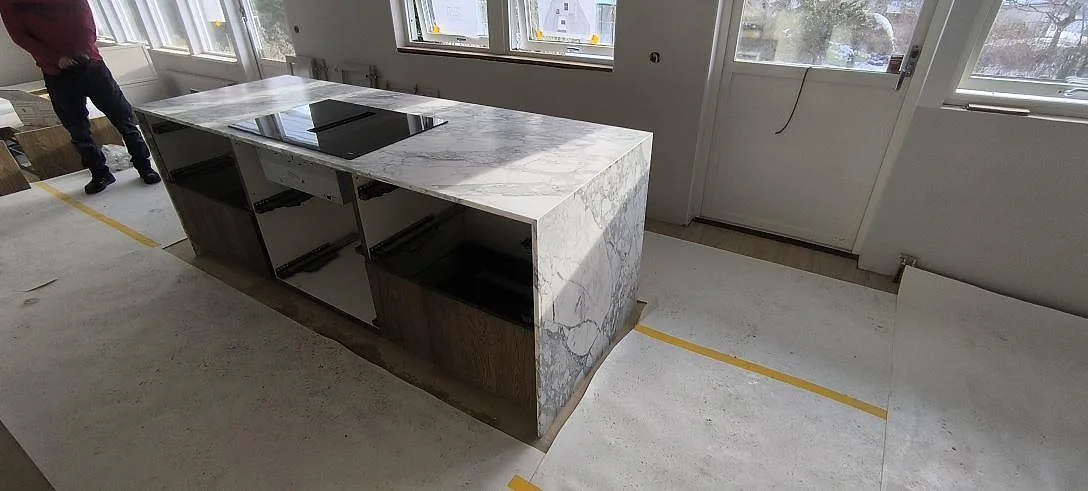 Köksö i vit Arabescato marmor med tydlig grå ådring och nedfälld gavel samt infälld induktionshäll, fotograferad under montering i ett kök i Stockholm.