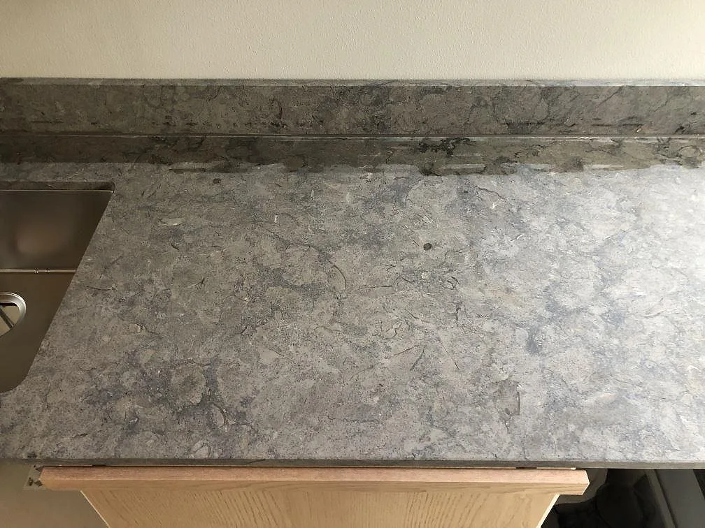 Köksbänkskiva i grå Azul Valverde-granit, matt yta, längs en köksvägg med ljusa eklaminerade överskåp, rostfri köksfläkt och underlimmad diskho i ett kök i Malmö.