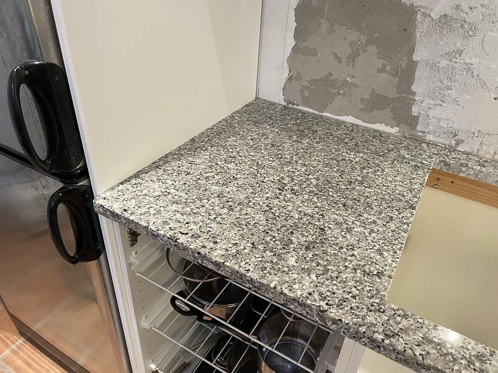 Sidobänk i Bianco Sardo granit, polerad, intill kyl och öppen köksförvaring under montering i Lund.
