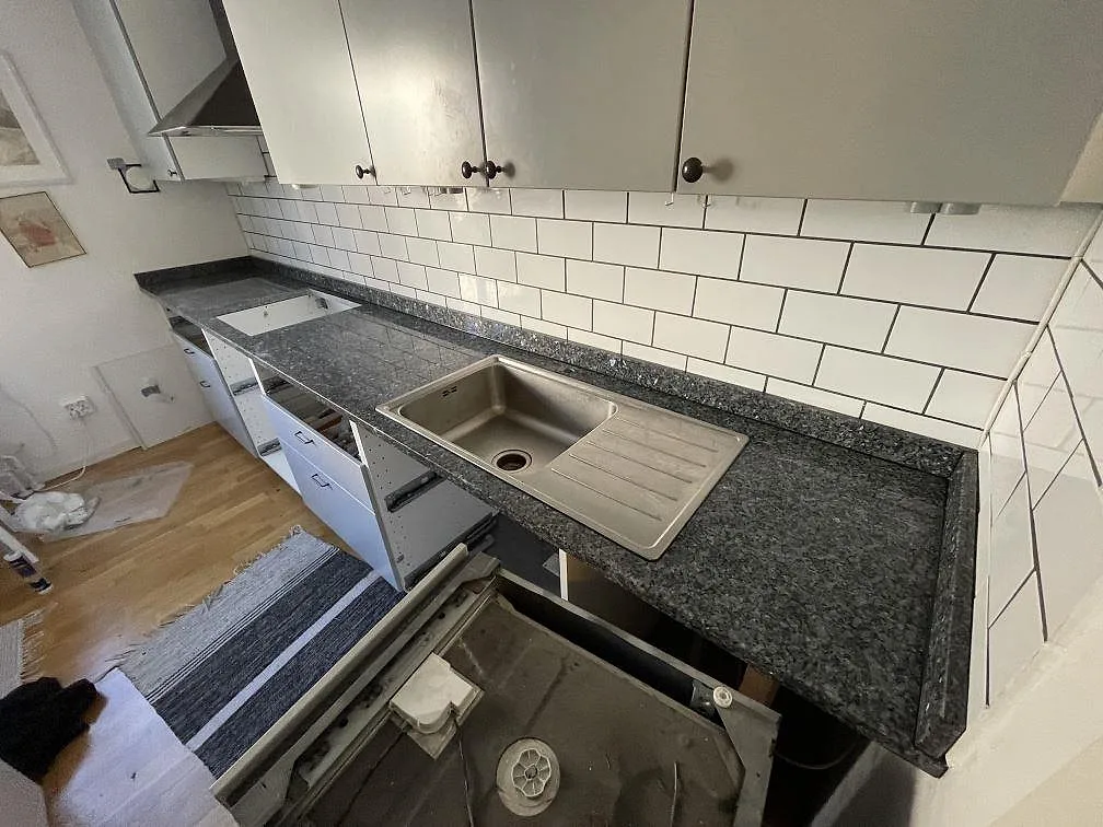 L-formad köksbänk i polerad Blue Pearl granit med rostfri diskho, hällurtag och vitt kakel i ett kök under montering i Sköndal.