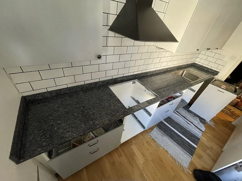 Köksbänkskiva i polerad Blue Pearl granit med uppvikt stänkkant, synligt hällurtag och rostfri diskho i ett kök i Sköndal.