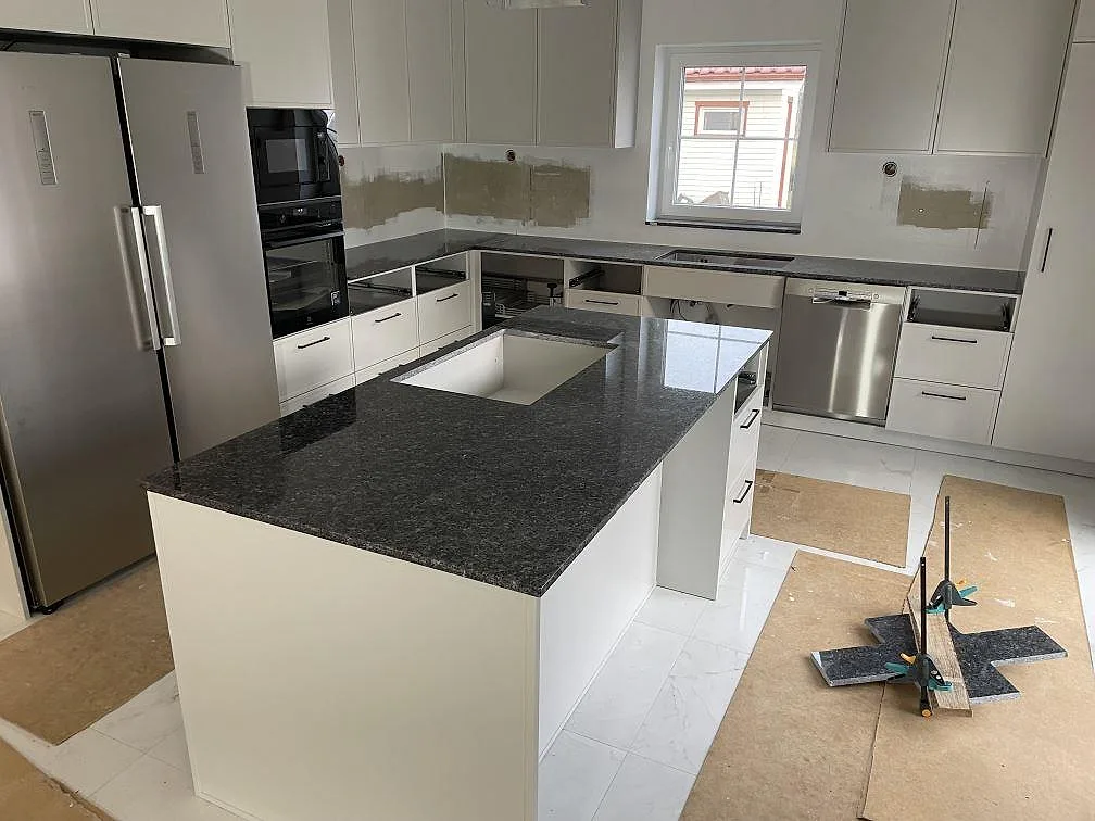 Köksbänkskiva i polerad Blue Pearl granit, 20 mm, med köksö och väggbänkar i ett kök under montering i Sölvesborg.