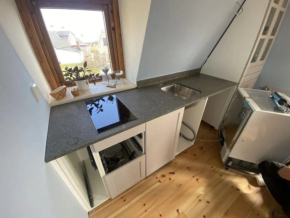 Köksbänkskiva i grå granit Bohus Grå, matt finish, längs fönstret i ett litet kök under snedtak med underlimmad rostfri diskho och induktionshäll i Brantevik.