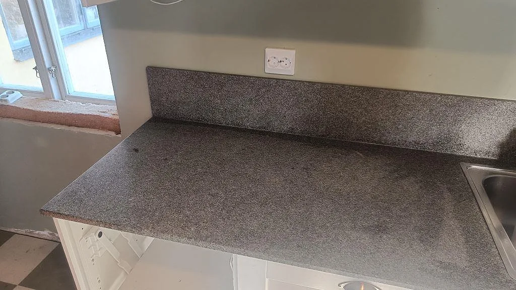 Närbild på köksbänkskiva i Bohus Grå granit med stänkkant, fönster och synlig diskbänkszon i Brantevik.