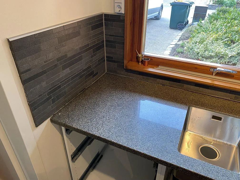 Hörnvy av köksbänkskiva i polerad Bohus Grå granit vid fönster, mörkt kakel och rostfri diskho i Höör.