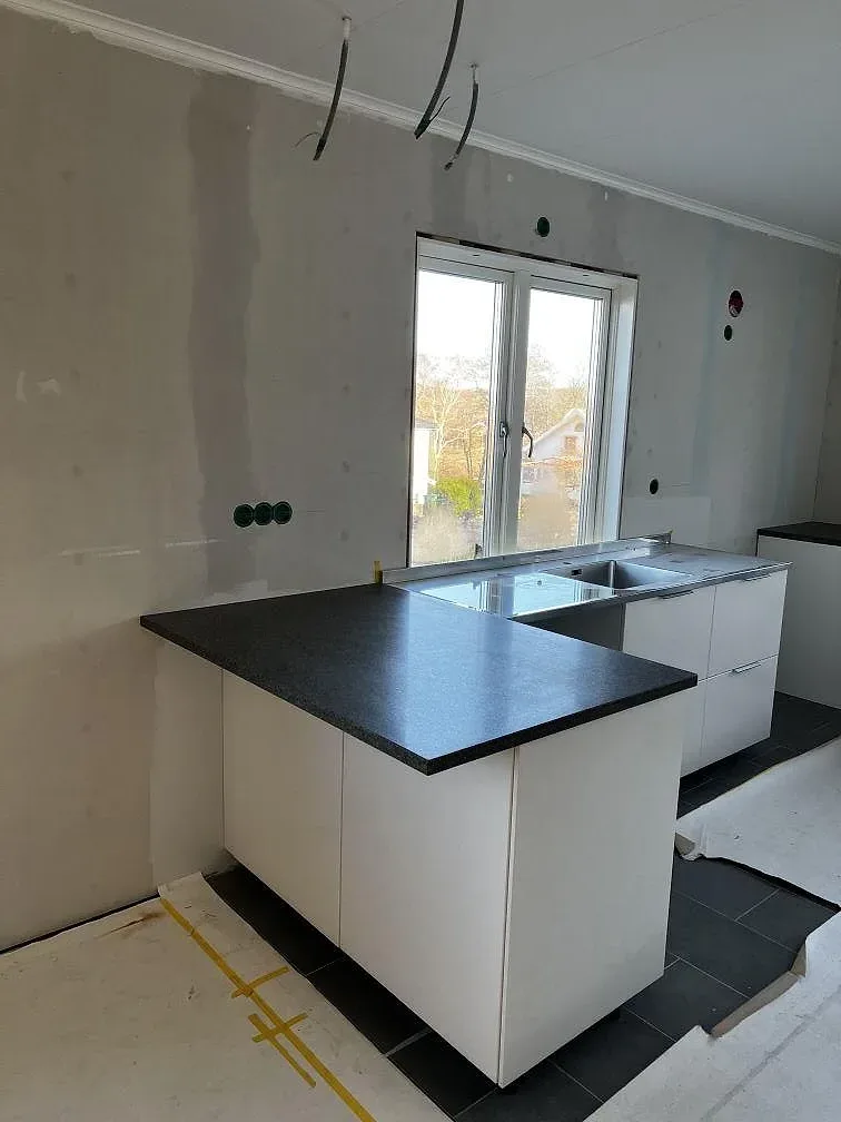 Köksbänkskiva och köksö i Bohus Grå granit, matt yta, i ett ljust kök under montering med vita stommar och stora fönster i Kungälv.