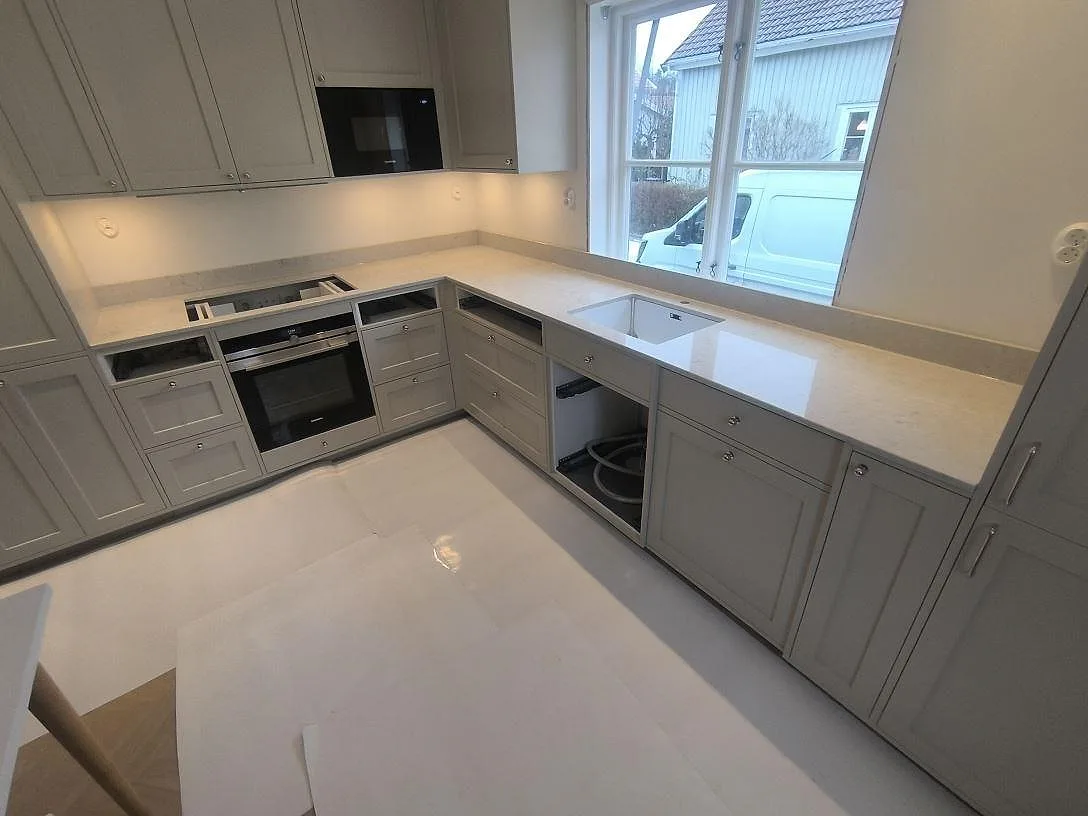 Köksbänkskiva i Caesarstone Bianco Drift, polerad kvartskomposit, med uppvikt stänkkant, diskho och hälluttag i ett kök i Enskede.