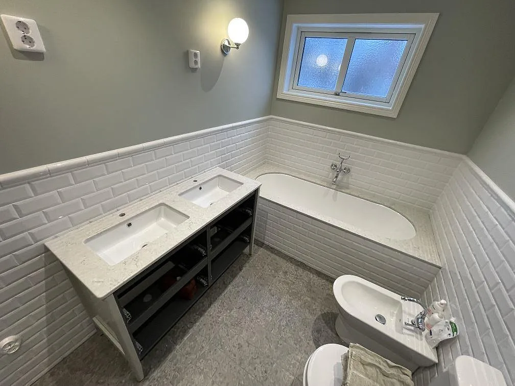 Bänkskiva i vit Caesarstone Bianco Drift, polerad, som tvättställsskiva med två underlimmade handfat på en mörk kommod och matchande stenram runt badkaret i ett badrum i Enskede.