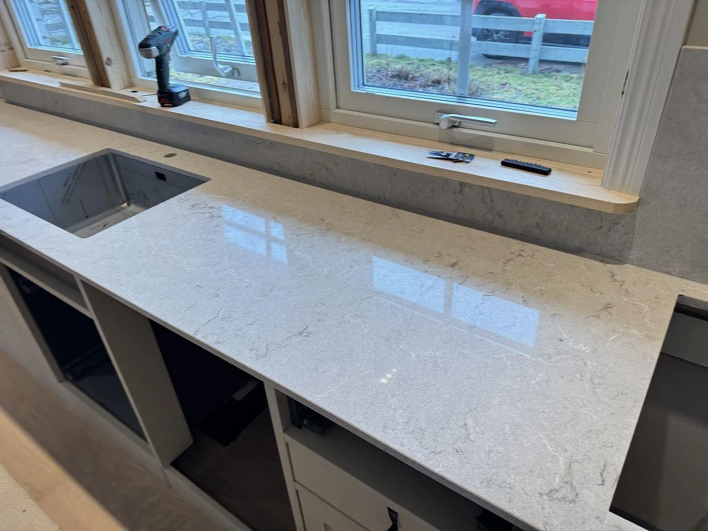 Köksbänkskiva i vit Caesarstone Bianco Drift, polerad kvartskomposit med grå ådring, med undermonterad rostfri diskho längs fönsterraden i ett kök i Kalmar.