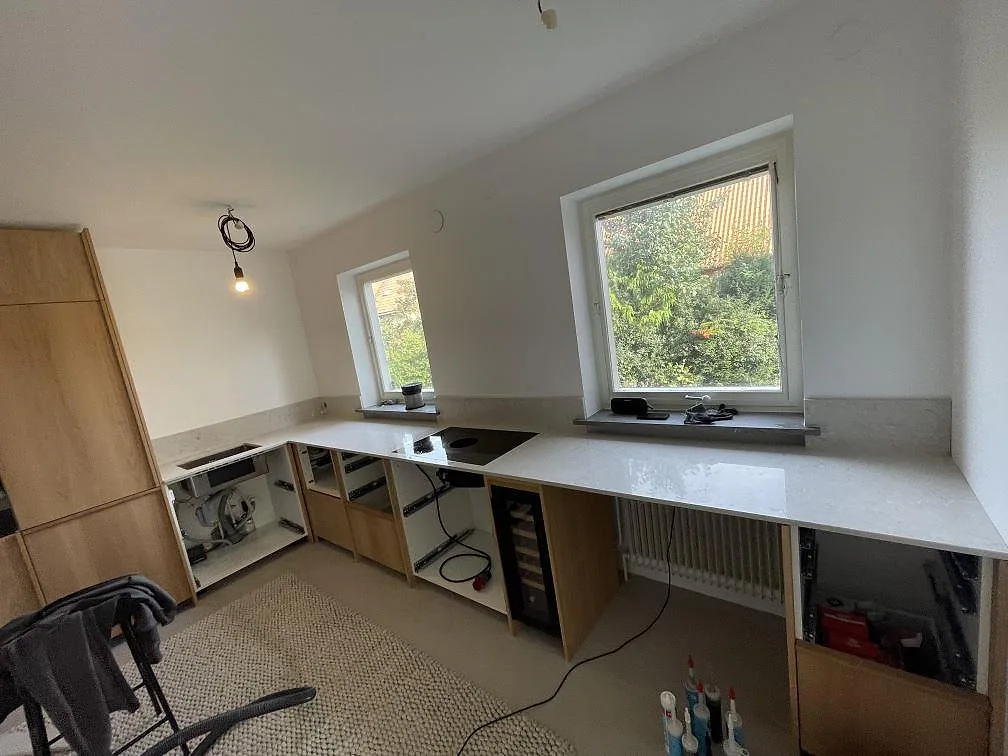 U-format kök i Malmö med polerad köksbänkskiva i Caesarstone Bianco Drift, svart induktionshäll, uppvikt stänkkant och trästommar under montering.
