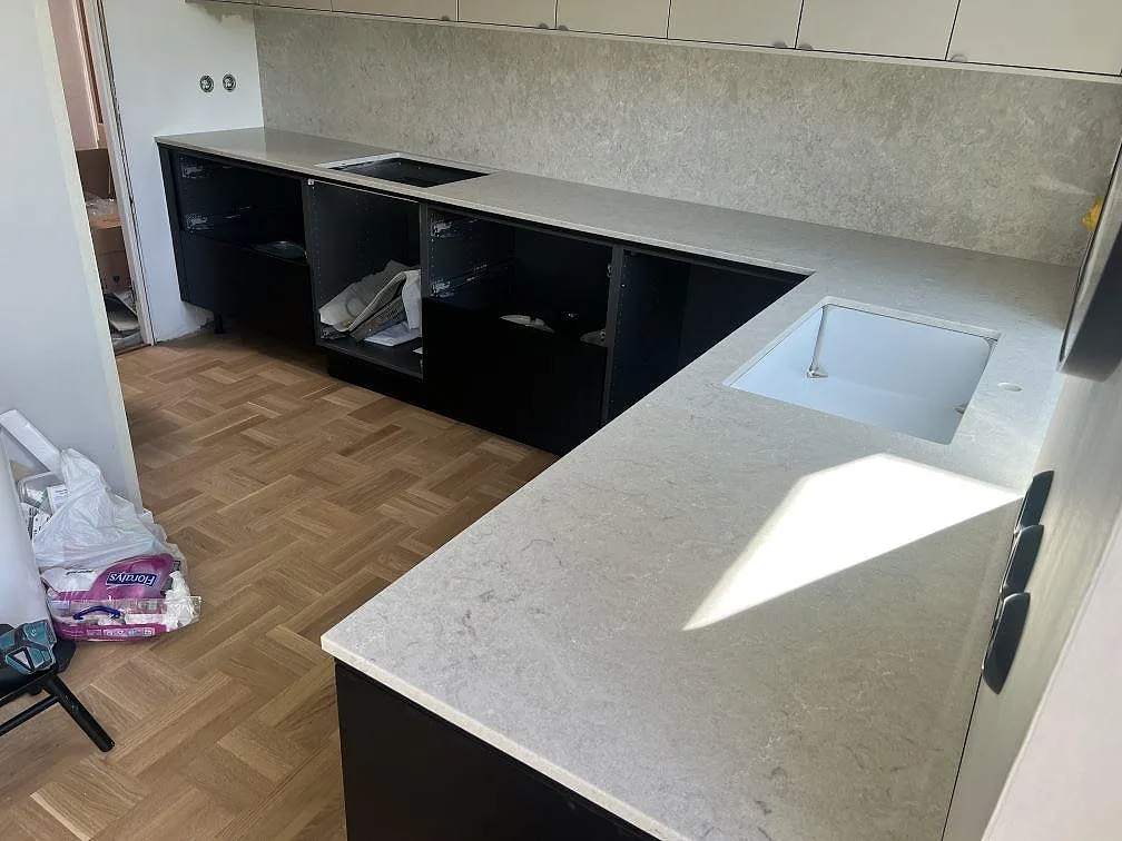 Polerad kompositbänkskiva i Caesarstone Bianco Drift med matchande stänkskydd, vit diskho och öppna underskåp under montering i Oxie.