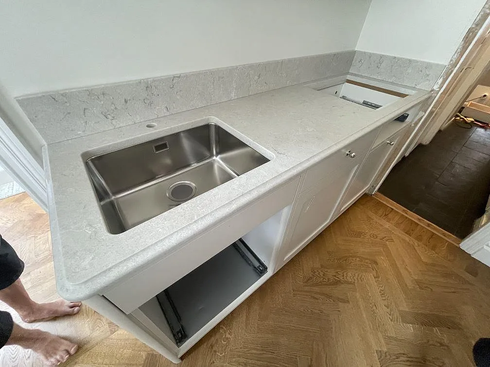 Köksbänkskiva i Caesarstone Bianco Drift, polerad vit kvartskomposit, med underlimmad rostfri diskho, hälluttag och uppvikt stänkkant i ett kök i Stockholm.