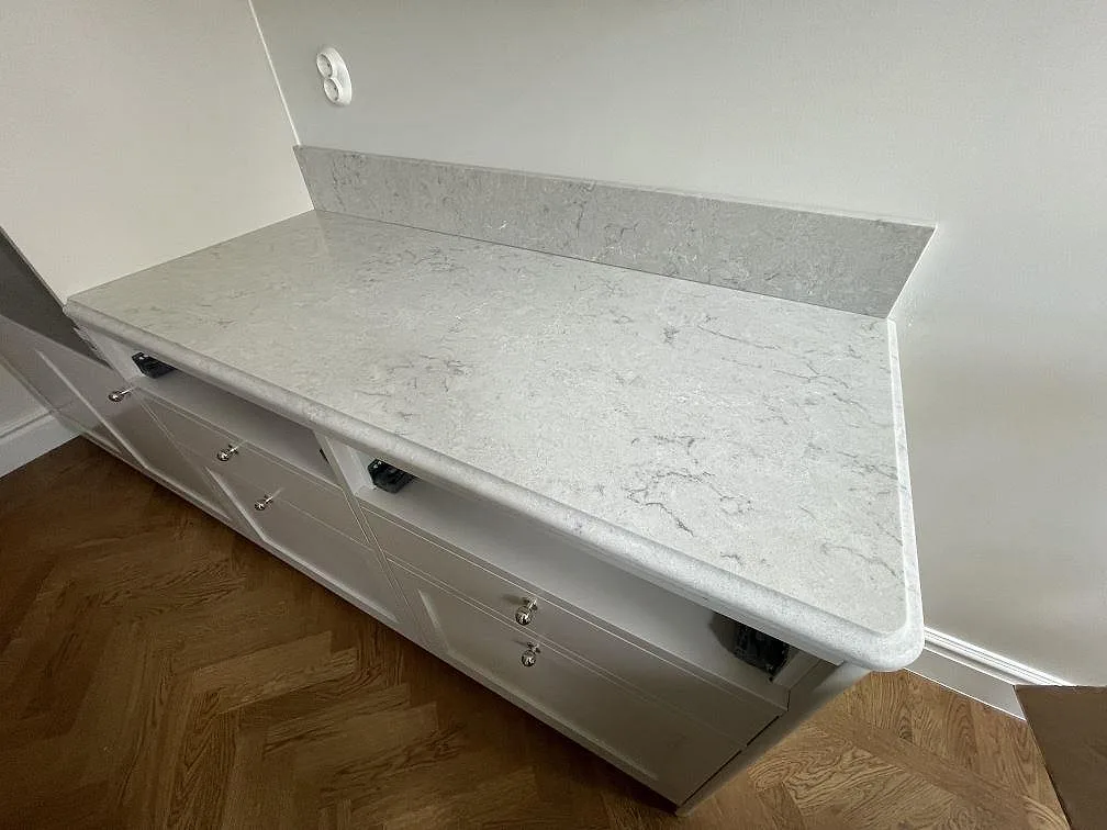 Polerad bänkskiva i Caesarstone Bianco Drift med mjuk grå ådring och uppvikt stänkkant på vita köksstommar i Stockholm.